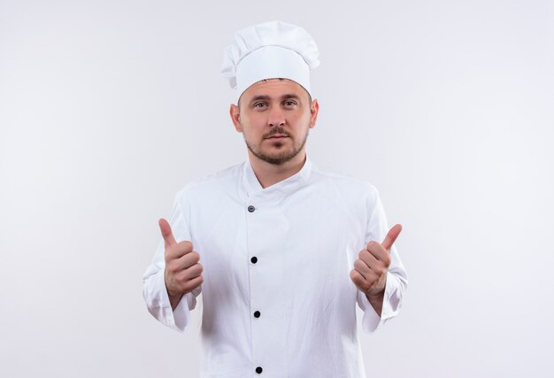 Confiant jeune beau cuisinier en uniforme de chef montrant les pouces vers le haut isolé sur mur blanc