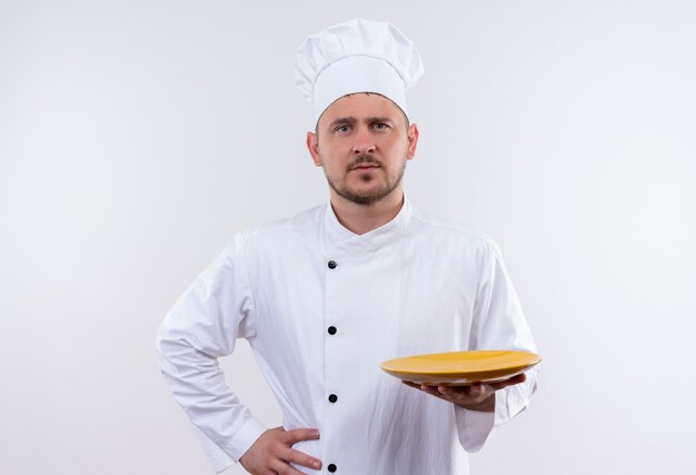 Confiant jeune beau cuisinier en uniforme de chef tenant une assiette avec une autre main sur la taille sur un mur blanc isolé