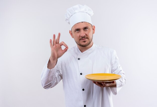 Confiant jeune beau cuisinier en uniforme de chef tenant une assiette et faisant signe ok sur un mur blanc isolé