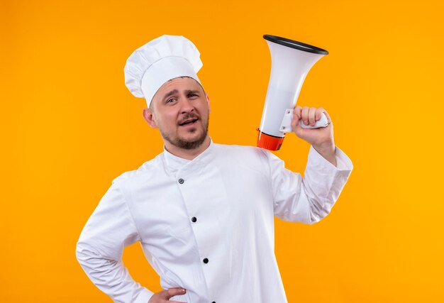 Confiant jeune beau cuisinier en uniforme de chef tenant le haut-parleur sur un mur orange isolé