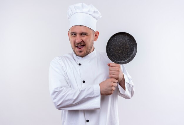 Confiant jeune beau cuisinier en uniforme de chef tenant une poêle à frire sur un mur blanc isolé