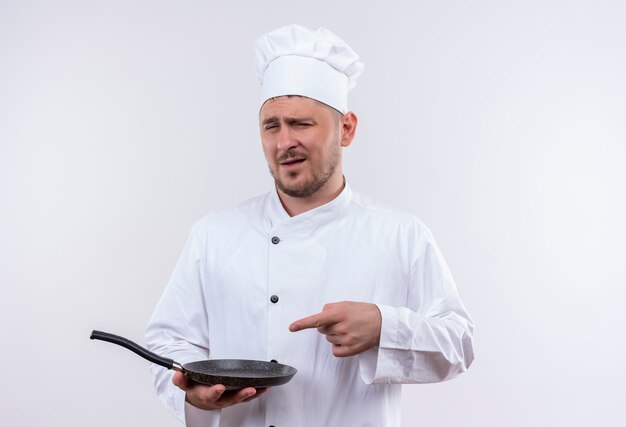 Confiant jeune beau cuisinier en uniforme de chef tenant et pointant sur une poêle à frire isolée sur mur blanc