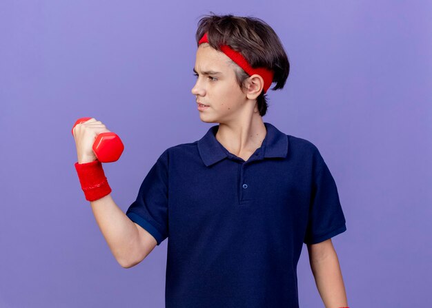 Confiant jeune beau garçon sportif portant un bandeau et des bracelets avec un appareil dentaire tenant et regardant haltère isolé sur mur violet
