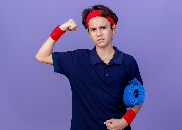 Confiant jeune beau garçon sportif portant un bandeau et des bracelets avec un appareil dentaire tenant un tapis de yoga à l'avant faisant un geste fort isolé sur un mur violet