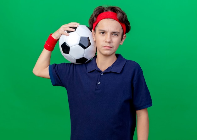 Confiant jeune beau garçon sportif portant un bandeau et des bracelets avec des appareils dentaires tenant un ballon de football sur l'épaule à l'avant isolé sur un mur vert