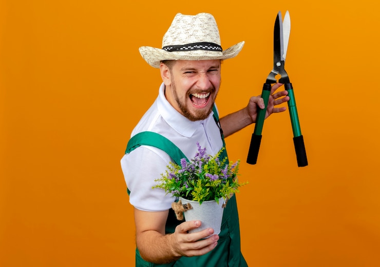 Photo gratuit confiant jeune beau jardinier slave en uniforme et chapeau debout en vue de profil tenant une plante à fleurs et sécateurs à crier isolé