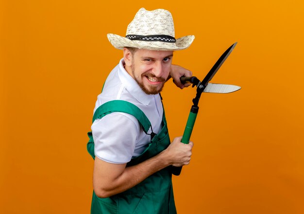 Confiant jeune beau jardinier slave en uniforme et chapeau debout en vue de profil tenant des sécateurs à isolé