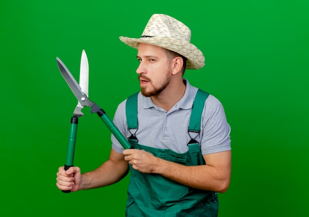 Photo gratuite confiant jeune beau jardinier slave en uniforme et chapeau à la tenue de sécateurs