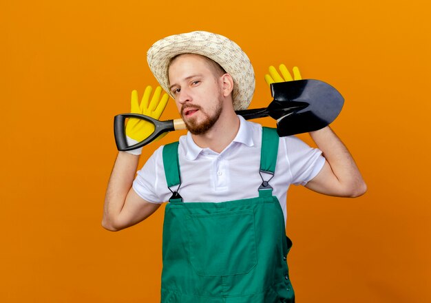 Confiant jeune beau jardinier slave en uniforme portant un chapeau et des gants de jardinage tenant une bêche derrière le cou isolé sur un mur orange avec espace de copie