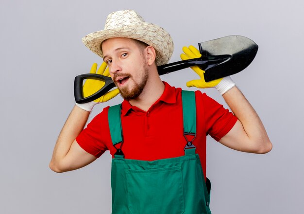 Confiant jeune beau jardinier slave en uniforme portant des gants de jardinage et un chapeau tenant une bêche derrière le cou isolé sur un mur blanc