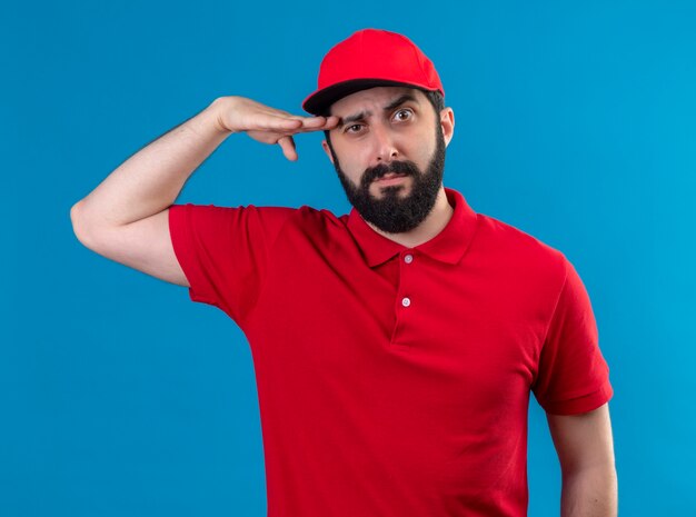 Confiant jeune beau livreur de race blanche portant l'uniforme rouge et une casquette mettant la main près de la tête isolée sur bleu