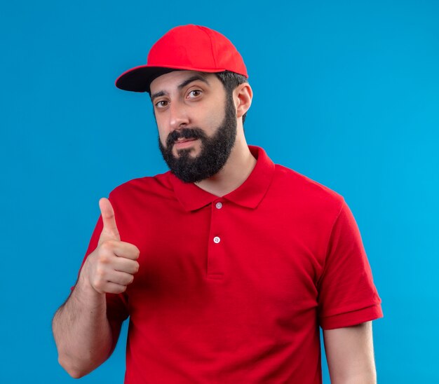 Confiant jeune beau livreur de race blanche portant l'uniforme rouge et une casquette montrant le pouce vers le haut isolé sur bleu