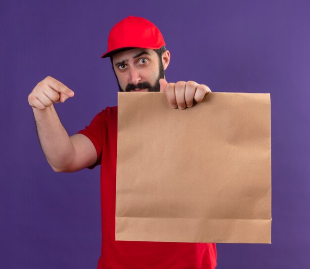 Confiant jeune beau livreur de race blanche portant l'uniforme rouge et une casquette s'étendant sur le paquet de papier vers la caméra et pointant vers elle isolé sur violet