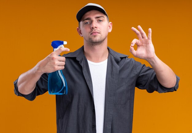 Confiant jeune beau mec de nettoyage portant un t-shirt et une casquette tenant un flacon pulvérisateur et montrant un geste correct isolé sur un mur orange