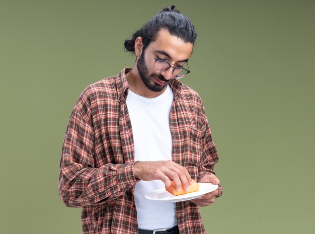 Confiant jeune beau mec de nettoyage portant t-shirt à laver la vaisselle avec une éponge isolé sur mur vert olive