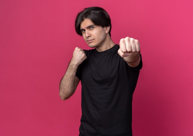 Confiant jeune beau mec portant un t-shirt noir debout dans la pose de combat isolé sur un mur rose avec espace de copie