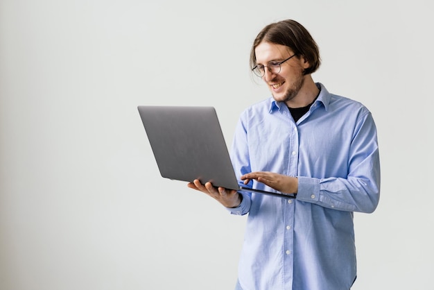 Confiant jeune bel homme en chemise tenant un ordinateur portable et souriant en se tenant debout sur fond blanc