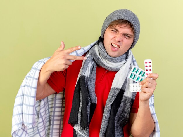 Confiant jeune bel homme malade blonde portant un chapeau d'hiver et une écharpe enveloppée dans un plaid tenant des paquets de pilules médicales regardant la caméra isolée sur fond vert olive