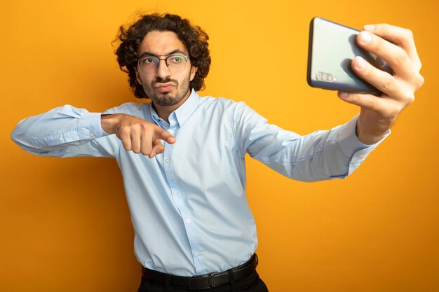 Confiant jeune bel homme portant des lunettes prenant selfie pointant sur téléphone isolé sur mur orange