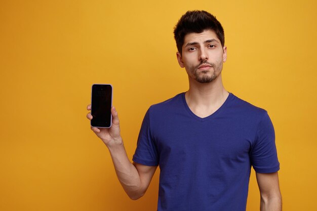 Confiant jeune bel homme regardant la caméra montrant un téléphone portable sur fond jaune