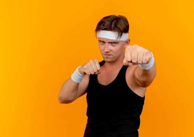 Confiant jeune bel homme sportif portant bandeau et bracelets faisant le geste de boxe isolé sur orange avec copie espace