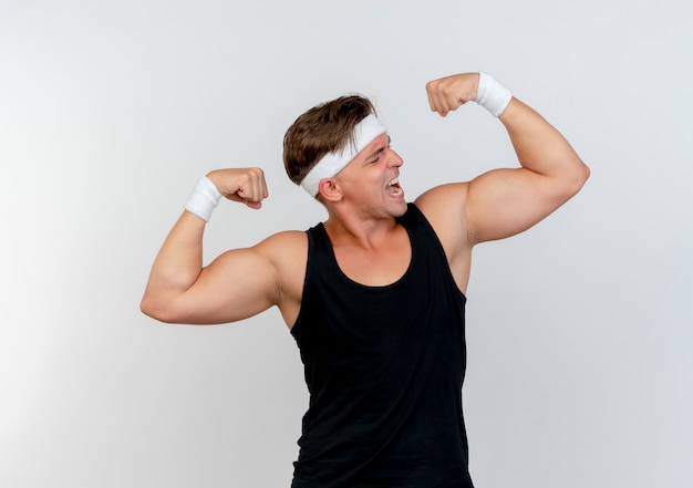 Confiant jeune bel homme sportif portant un bandeau et des bracelets faisant des gestes forts et en regardant ses muscles isolés sur blanc