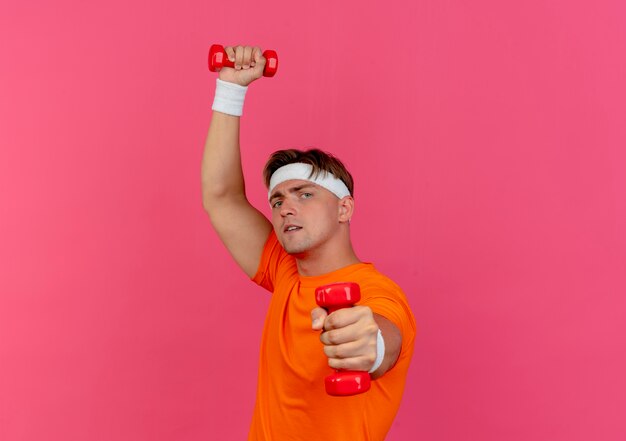Confiant jeune bel homme sportif portant bandeau et bracelets soulevant et étirant des haltères isolés sur rose avec espace copie