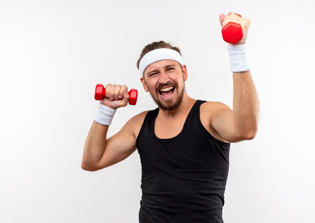 Confiant jeune bel homme sportif portant un bandeau et des bracelets soulevant des haltères isolés sur un mur blanc avec un espace de copie