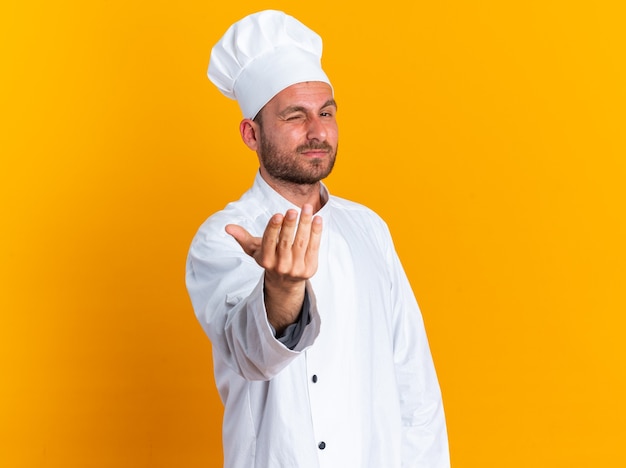 Confiant jeune cuisinier masculin caucasien en uniforme de chef et casquette regardant la caméra en faisant un clin d'œil faisant venir ici geste isolé sur mur orange avec espace de copie