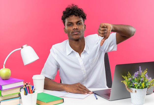 Confiant jeune étudiant afro-américain assis au bureau avec des outils scolaires pouce vers le bas isolé sur mur rose