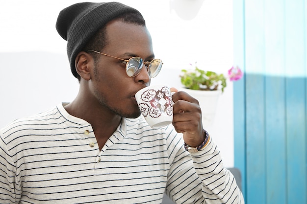 Confiant jeune étudiant afro-américain habillé avec style en appréciant le café au café du collège. Homme à la peau sombre à la mode avec une tasse de boire du thé tout en déjeunant au restaurant confortable seul