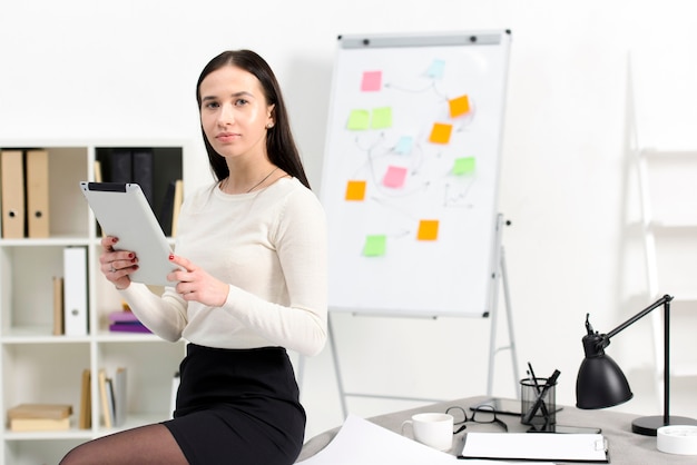 Confiant jeune femme d&#39;affaires tenant une tablette numérique à la main, assis sur la table sur le lieu de travail