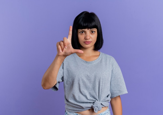 Confiant jeune femme brune caucasienne gestes signe de main pistolet isolé sur fond violet avec espace copie
