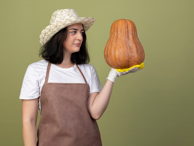 Confiant jeune femme brune jardinière en uniforme portant chapeau de jardinage et des gants tient et regarde la citrouille isolée sur mur vert olive