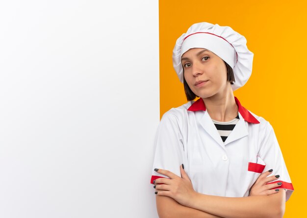 Confiant jeune femme cuisinier en uniforme de chef debout avec une posture fermée devant un mur blanc isolé sur orange avec copie espace