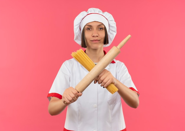 Confiant jeune femme cuisinier en uniforme de chef gestes non avec des pâtes spaghetti et rouleau à pâtisserie isolé sur rose avec copie espace