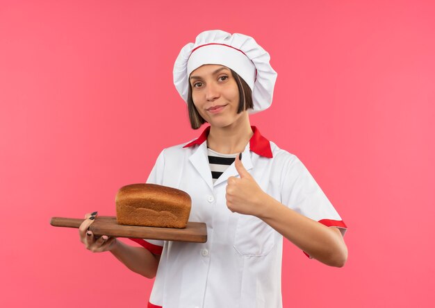 Confiant jeune femme cuisinier en uniforme de chef tenant une planche à découper avec du pain dessus et montrant le pouce vers le haut isolé sur rose