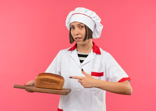 Confiant jeune femme cuisinier en uniforme de chef tenant et pointant sur une planche à découper avec du pain dessus isolé sur rose