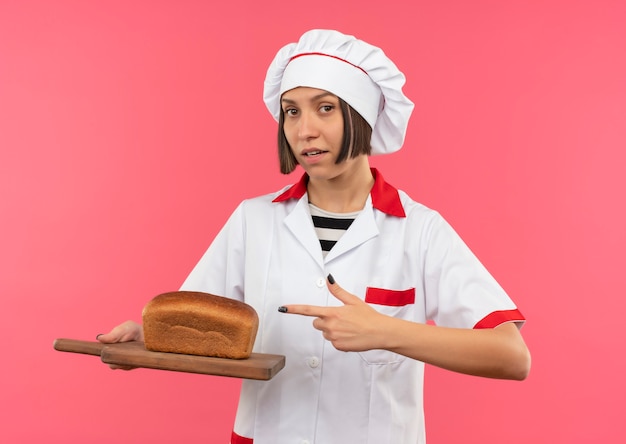 Confiant jeune femme cuisinier en uniforme de chef tenant et pointant sur une planche à découper avec du pain dessus isolé sur rose