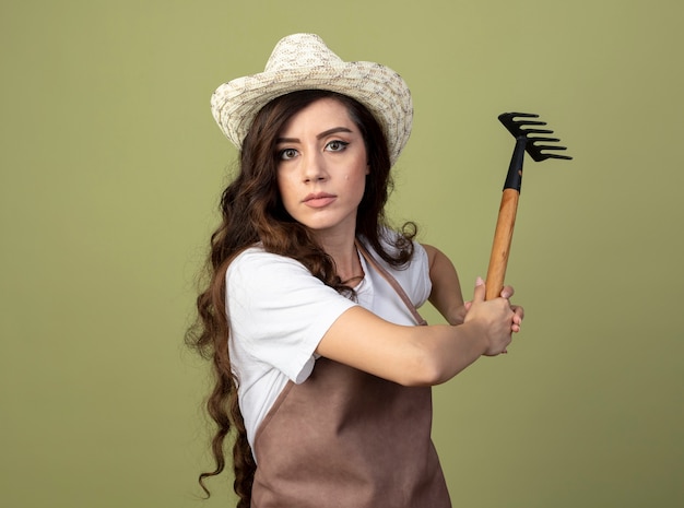 Confiant jeune femme jardinier en uniforme portant chapeau de jardinage tenant râteau isolé sur mur vert olive