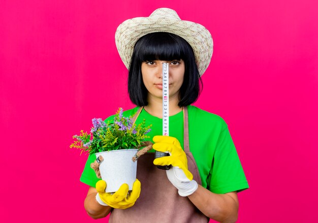 Photo gratuite confiant jeune femme jardinière en uniforme portant chapeau de jardinage détient pot de fleurs et ruban à mesurer