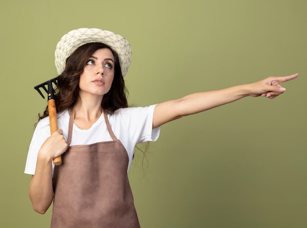 Confiant jeune femme jardinière en uniforme portant chapeau de jardinage détient râteau et points sur le côté isolé sur mur vert olive