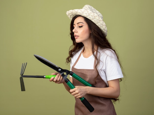 Photo gratuite confiant jeune femme jardinière en uniforme portant chapeau de jardinage détient tondeuses et houe râteau isolé sur mur vert olive