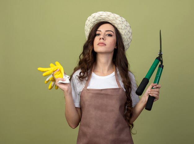 Confiant jeune femme jardinière en uniforme portant chapeau de jardinage détient des tondeuses de jardin et des gants isolés sur mur vert olive