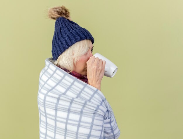 Confiant jeune femme malade blonde portant un chapeau d'hiver et une écharpe enveloppée dans des stands à carreaux sur le côté buvant de tasse isolée sur mur vert olive