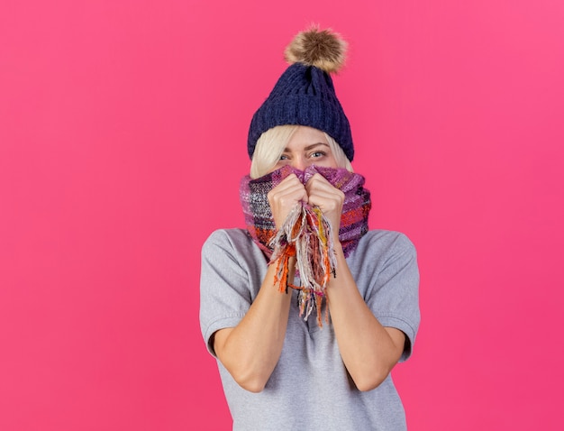 Confiant jeune femme malade blonde portant un chapeau d'hiver et tient un foulard isolé sur un mur rose