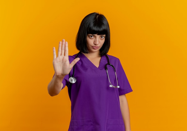 Confiant jeune femme médecin brune en uniforme avec des gestes stéthoscope signe de la main d'arrêt