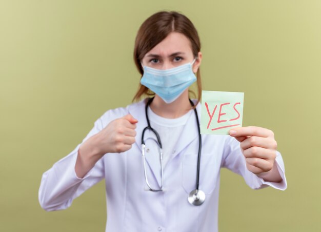 Confiant jeune femme médecin portant une robe médicale, un masque et un stéthoscope qui s'étend oui note avec le poing fermé sur un espace vert isolé