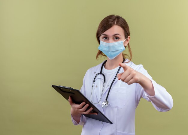 Confiant jeune femme médecin portant une robe médicale, un masque et un stéthoscope tenant le presse-papiers et pointant sur un espace vert isolé avec espace de copie