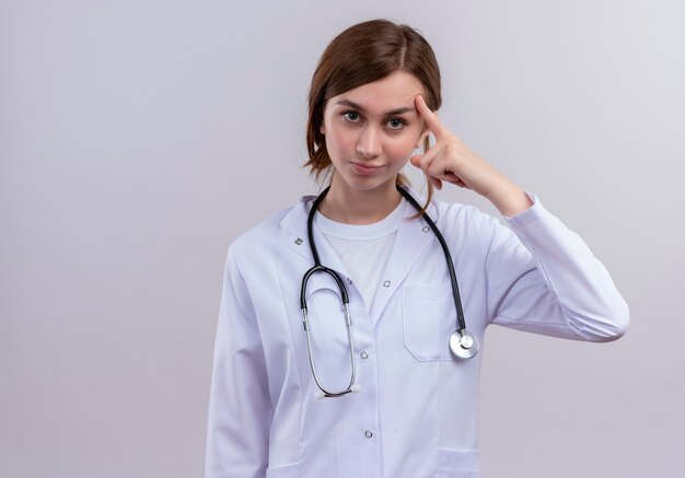 Confiant jeune femme médecin portant une robe médicale et un stéthoscope mettant le doigt sur le temple avec copie espace